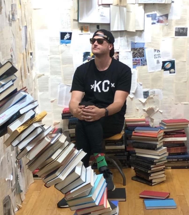 Adam seated among stacks of books and paper-covered walls.